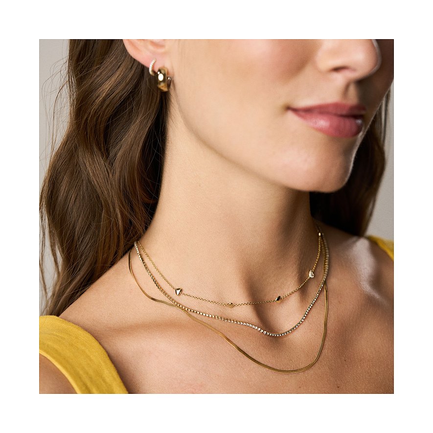 Gros plan d'une femme avec trois colliers Fossil dorés. Deux avec pendentifs cœur, un avec des perles de verre.