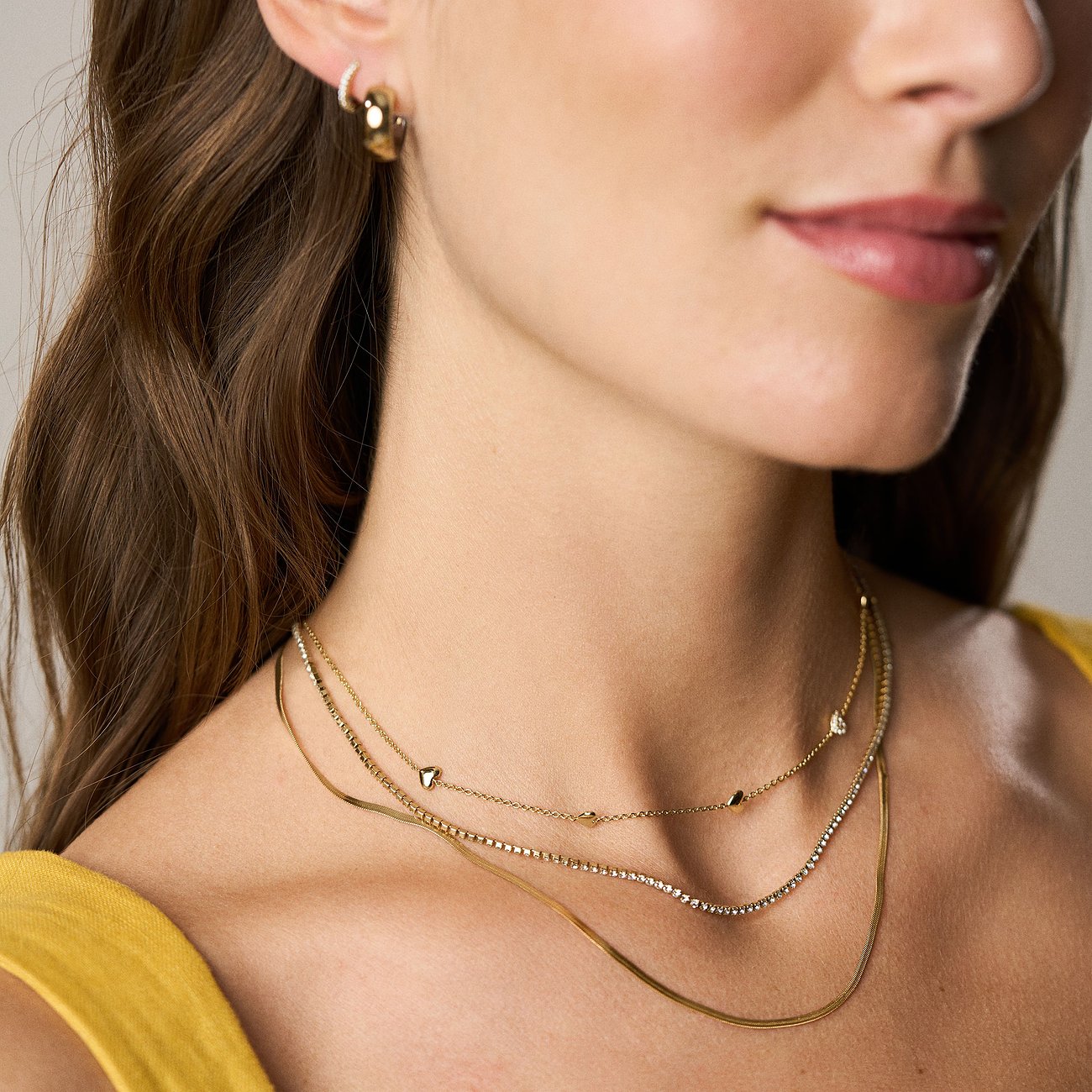Gros plan d'une femme avec trois colliers Fossil dorés. Deux avec pendentifs cœur, un avec des perles de verre.