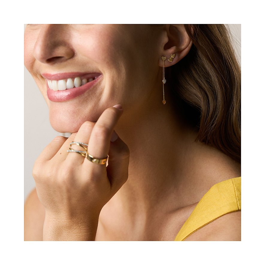 Nahaufnahme einer lächelnden Frau, die Fossil Hängeohrringe aus goldfarbenem Metall mit Herzmotiven und Glassteinen trägt.