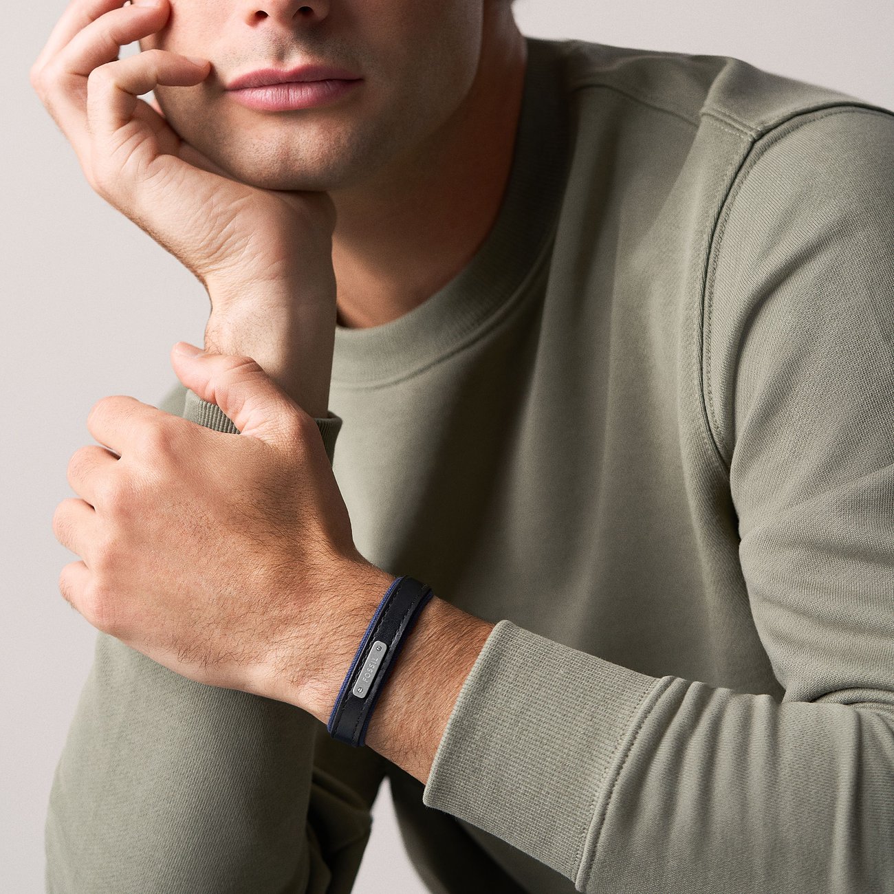 Un uomo in maglione grigio, mano sul mento, indossa un bracciale Fossil in pelle blu e metallo.