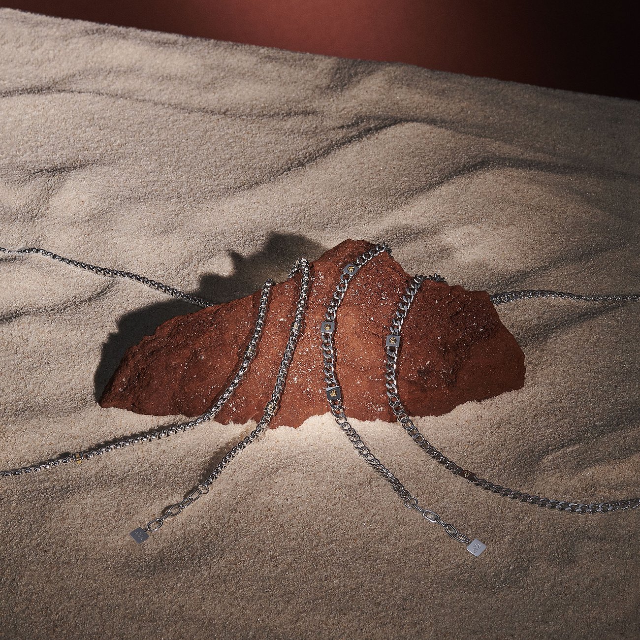 Collier Morellato en acier inoxydable, partiellement avec des éléments en or jaune 750, posé sur roche brune dans le sable.