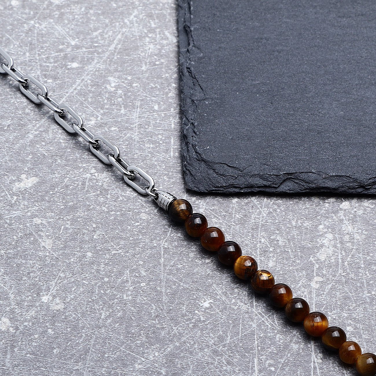 Morellato-armbandet i rostfritt stål och tigeröga, tillverkat av en kedja och bruna pärlor, ligger på grå bakgrund.