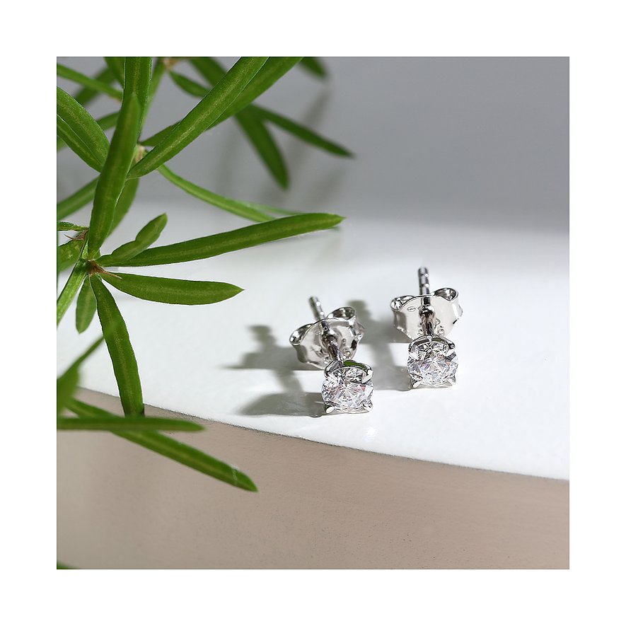 Ein Paar glänzende CHRIST Ohrstecker aus 585 Weißgold, besetzt mit je einem funkelnden Diamanten, daneben grüne Blätter.
