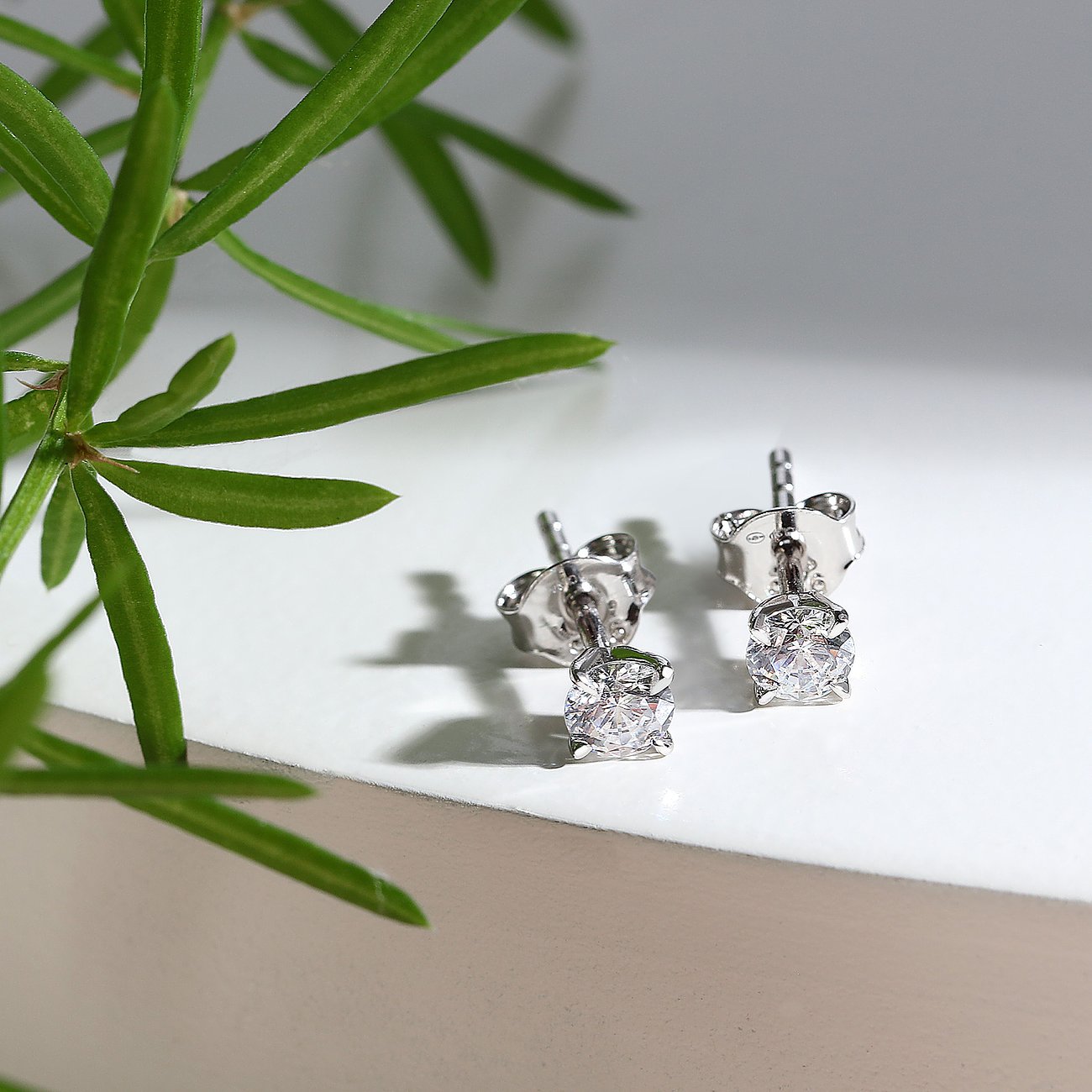 Ein Paar glänzende CHRIST Ohrstecker aus 585 Weißgold, besetzt mit je einem funkelnden Diamanten, daneben grüne Blätter.