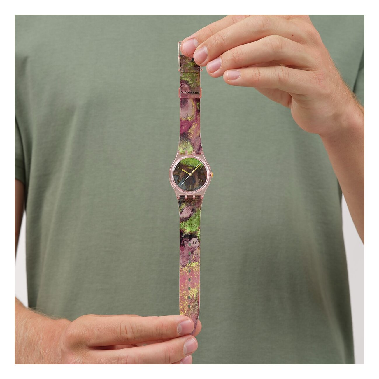 Ein Mann hält eine Swatch Unisexuhr aus Kunststoff mit einem bunten, gemusterten Armband und rosafarbenem Gehäuse.