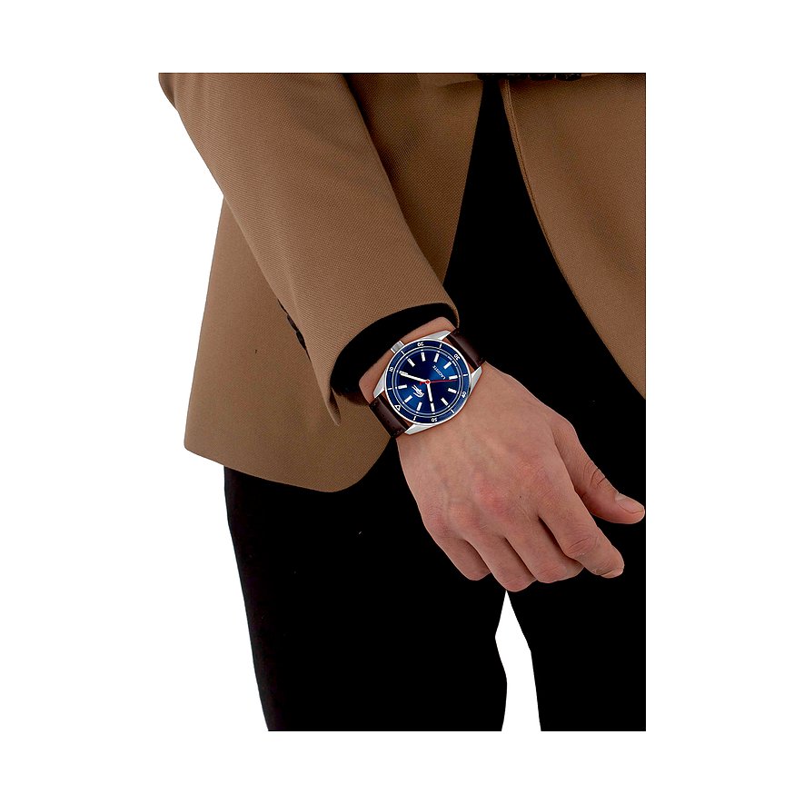 Eine Lacoste Herrenuhr aus Edelstahl mit blauem Zifferblatt und braunem Armband wird am Handgelenk getragen.