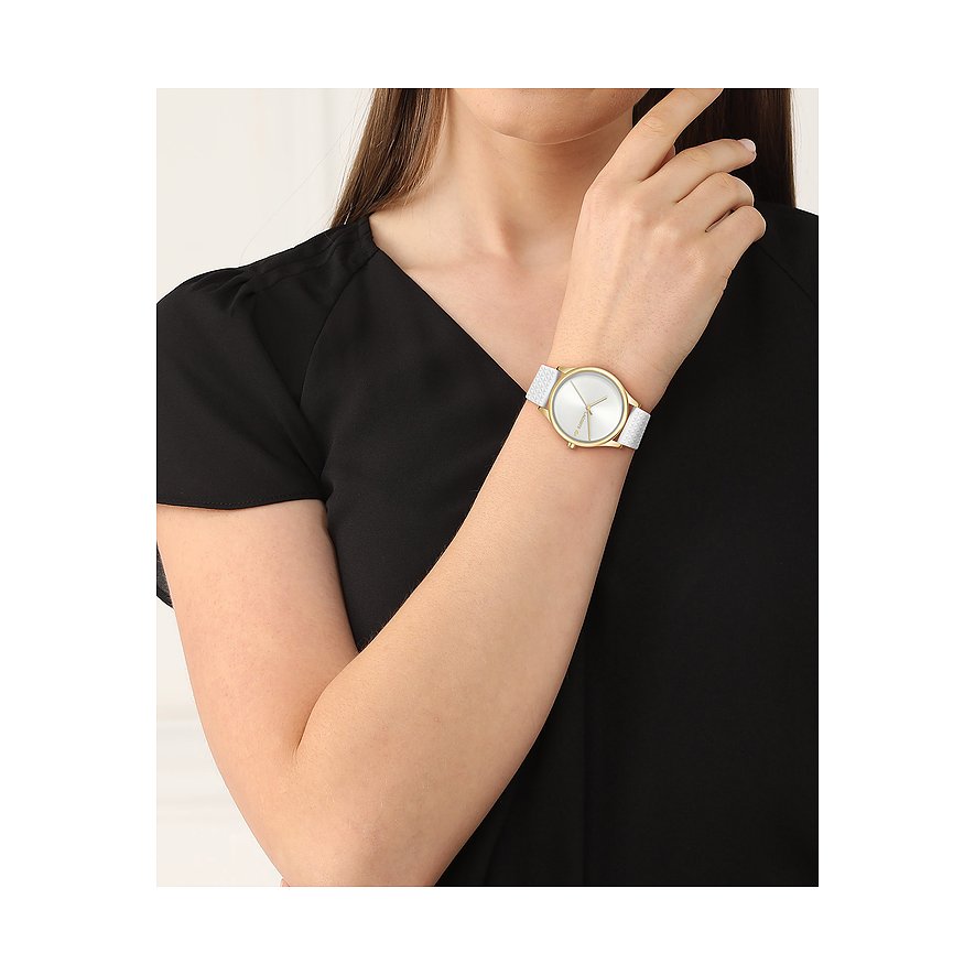 Am Handgelenk einer Frau ist eine Lacoste Damenuhr aus Edelstahl mit goldenem Gehäuse und weißem Armband zu sehen.