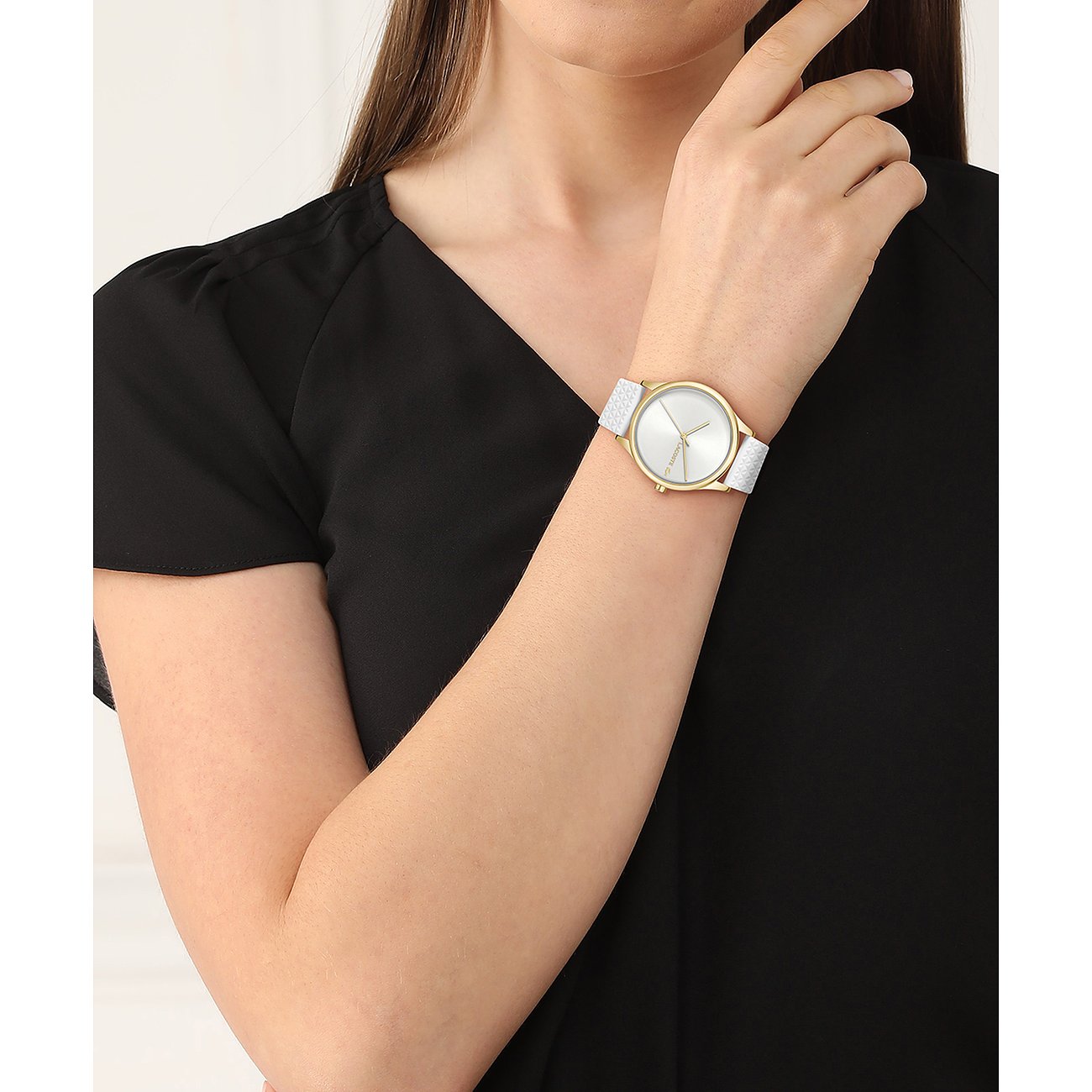 Am Handgelenk einer Frau ist eine Lacoste Damenuhr aus Edelstahl mit goldenem Gehäuse und weißem Armband zu sehen.