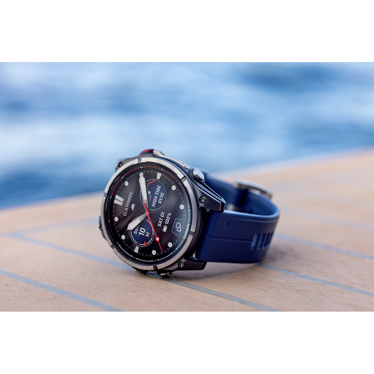 Nahaufnahme einer Garmin Smartwatch aus Metall mit dunkelblauem Armband, die auf einem Holzdeck liegt.
