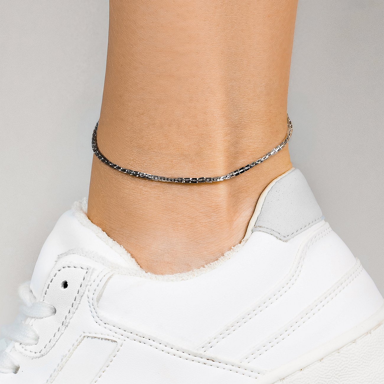 Ein am Knöchel getragenes, filigranes Copenhagen Studios Fußkettchen aus silberfarbenem Edelstahl über einem weißen Schuh.