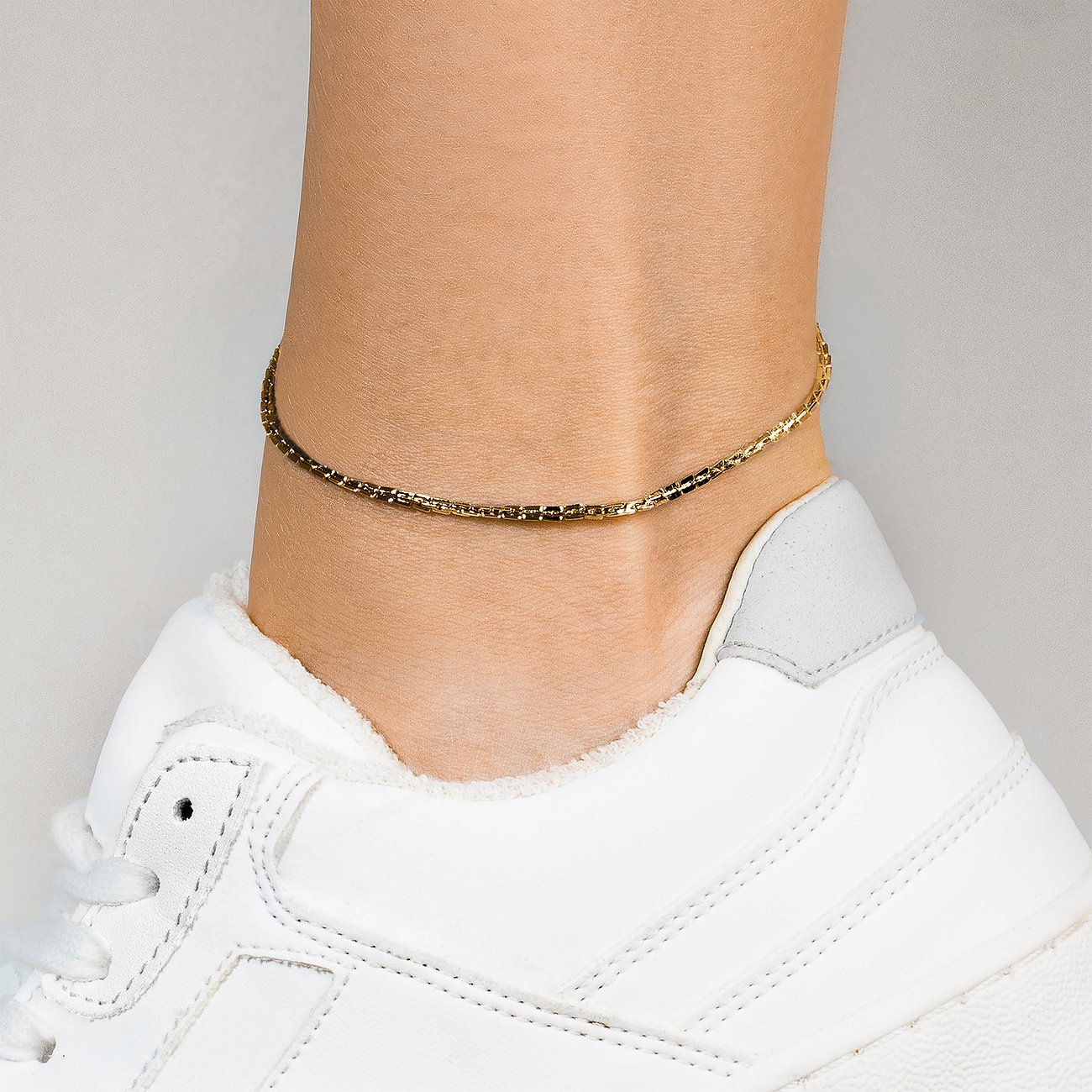 Copenhagen Studios Fußkette aus goldfarbenem Edelstahl, filigran, um einen Fußknöchel getragen, über einem weißen Sneaker.