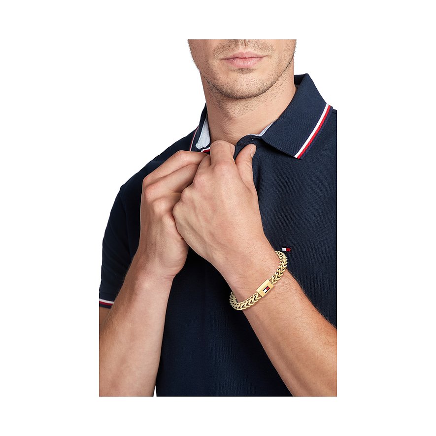 Ein goldenes Tommy Hilfiger Armband aus Edelstahl an einem Herrenarm, der ein dunkelblaues Poloshirt trägt.