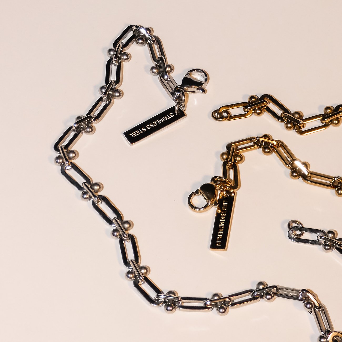 Zwei Liebeskind Armreifen aus Edelstahl, einer in Silber und der andere in Gold, beide mit Kettengliedern und kleinen Kugeln.
