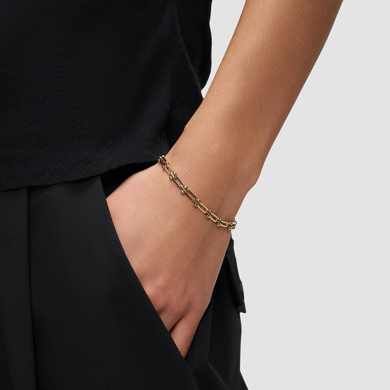Liebeskind armbånd af guldfarvet rustfrit stål båret på et models håndled, som rækker ned i en bukselomme.