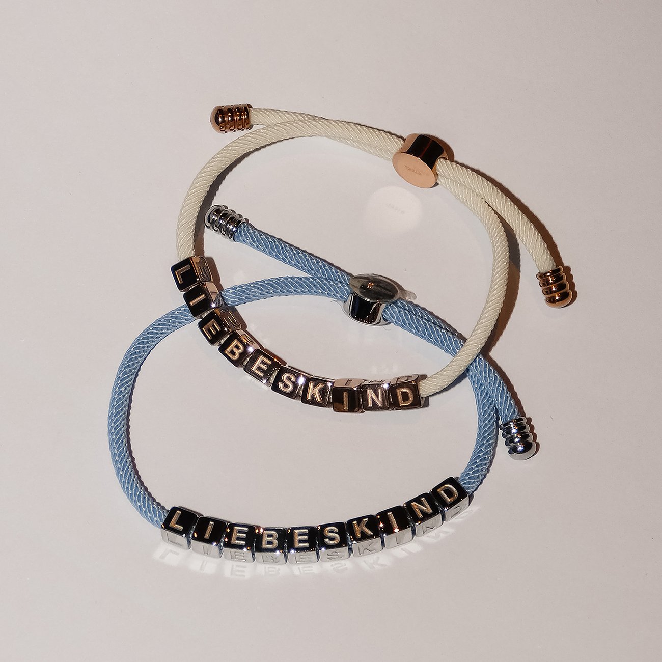 Zwei Liebeskind-Armbänder aus Edelstahl und Textil, eines hellblau und eines cremefarben, nebeneinander liegend.