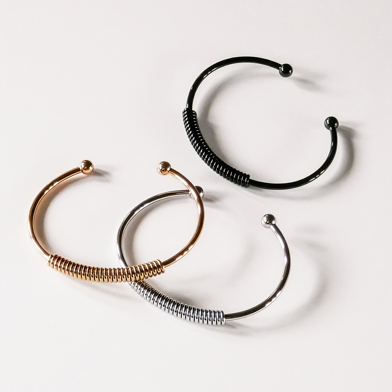Drei Liebeskind Edelstahl Armreifen in Schwarz, Roségold und Silber, jeweils mit Federspiraldetail und Kugelenden.