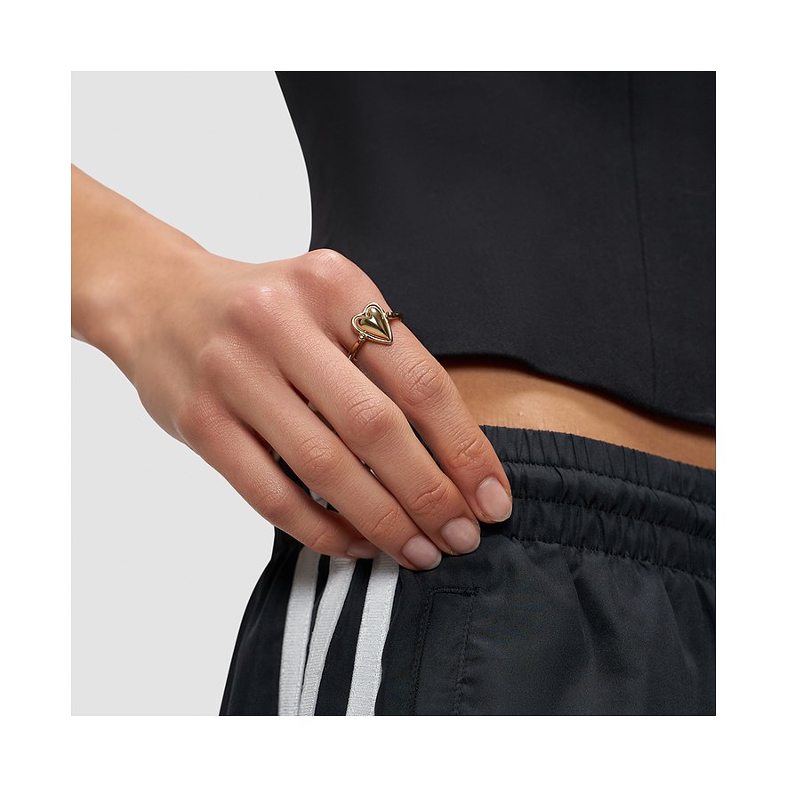 Goldfarbener Liebeskind Edelstahl Ring mit Herzmotiv am Ringfinger einer Person mit dunklem Oberteil und gestreifter Hose.