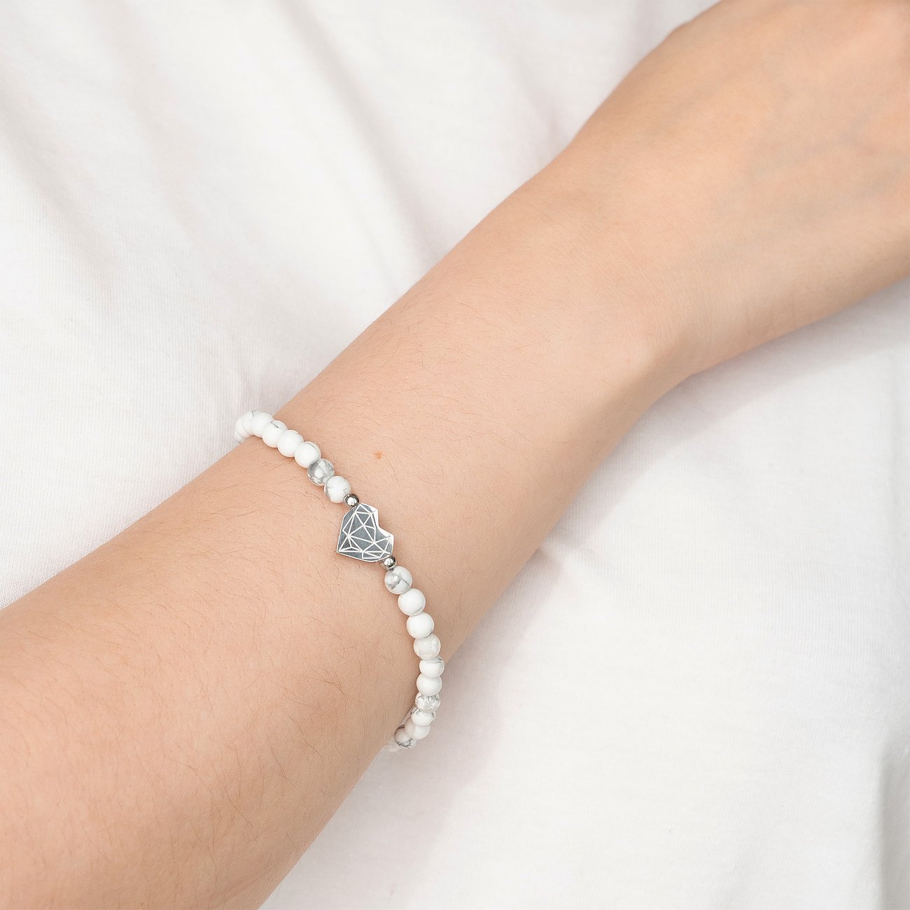 Ein weißes Perlenarmband der Marke Liebeskind mit einem herzförmigen Edelstahl-Anhänger wird an einem Arm getragen.