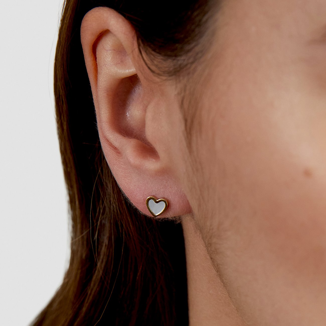 Gros plan : oreille droite avec petite boucle d'oreille cœur s.Oliver dorée en acier inoxydable. Centre du cœur blanc, nacré.