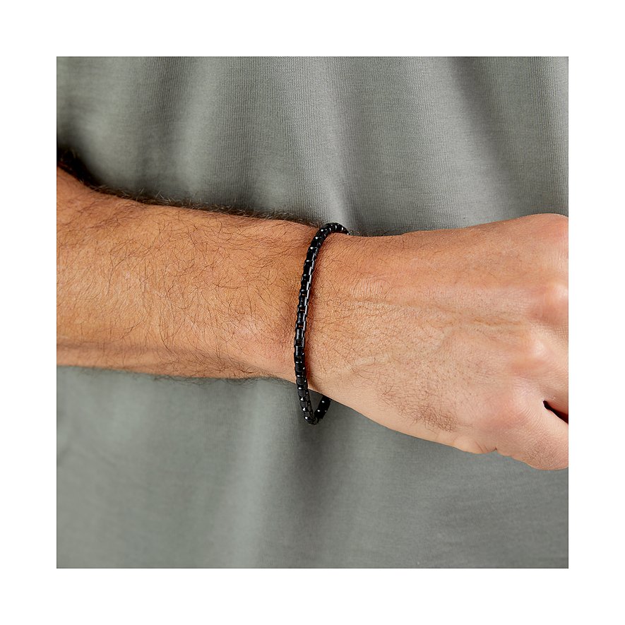 Ett s.Oliver-armband i svart rostfritt stål på en mans handled, framför en grågrön T-shirt.