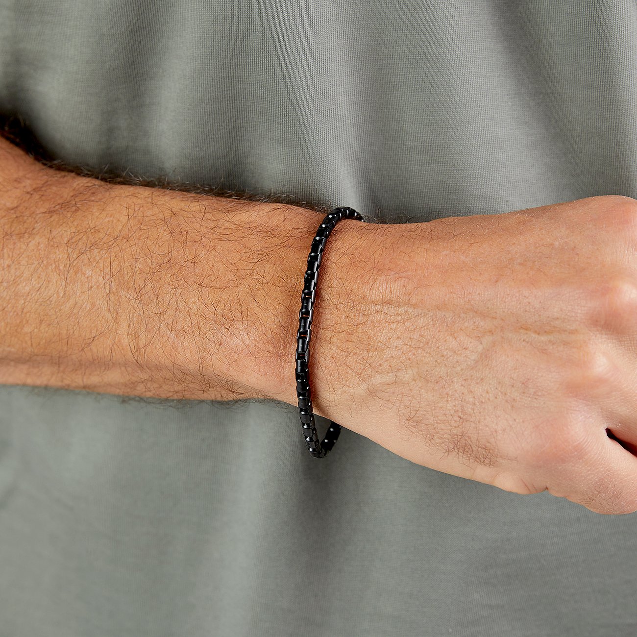Ett s.Oliver-armband i svart rostfritt stål på en mans handled, framför en grågrön T-shirt.