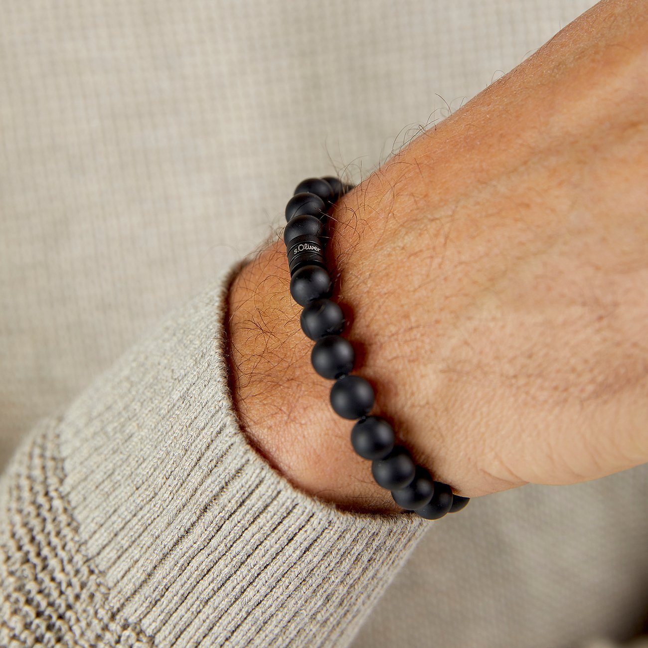 Un bracelet s.Oliver en pierres noires avec un pendentif de marque, porté au poignet d'un homme.