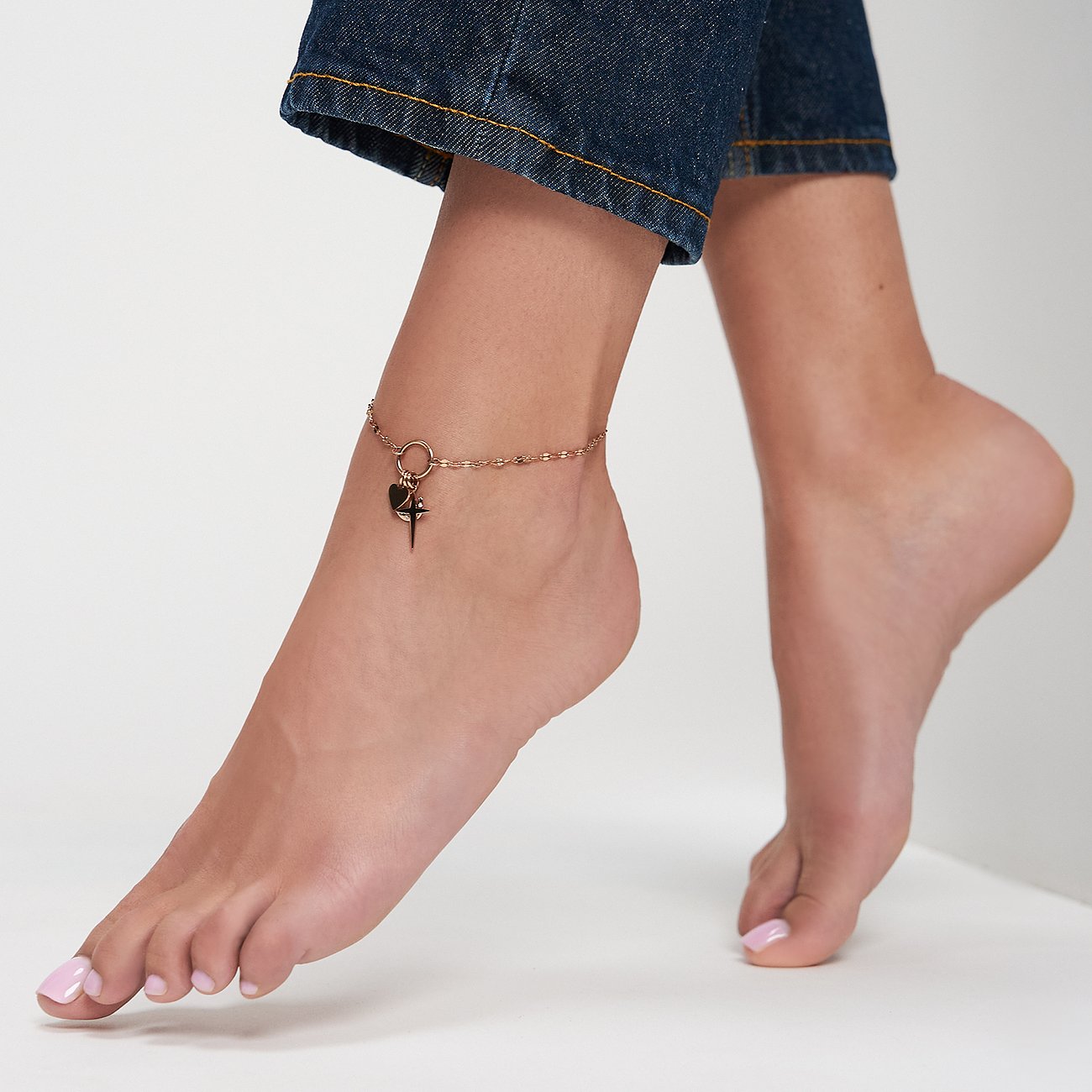 Am Fuß einer Person ist eine goldfarbene Tamaris Fußkette aus Edelstahl mit einem Herz- und Kreuzanhänger zu sehen.