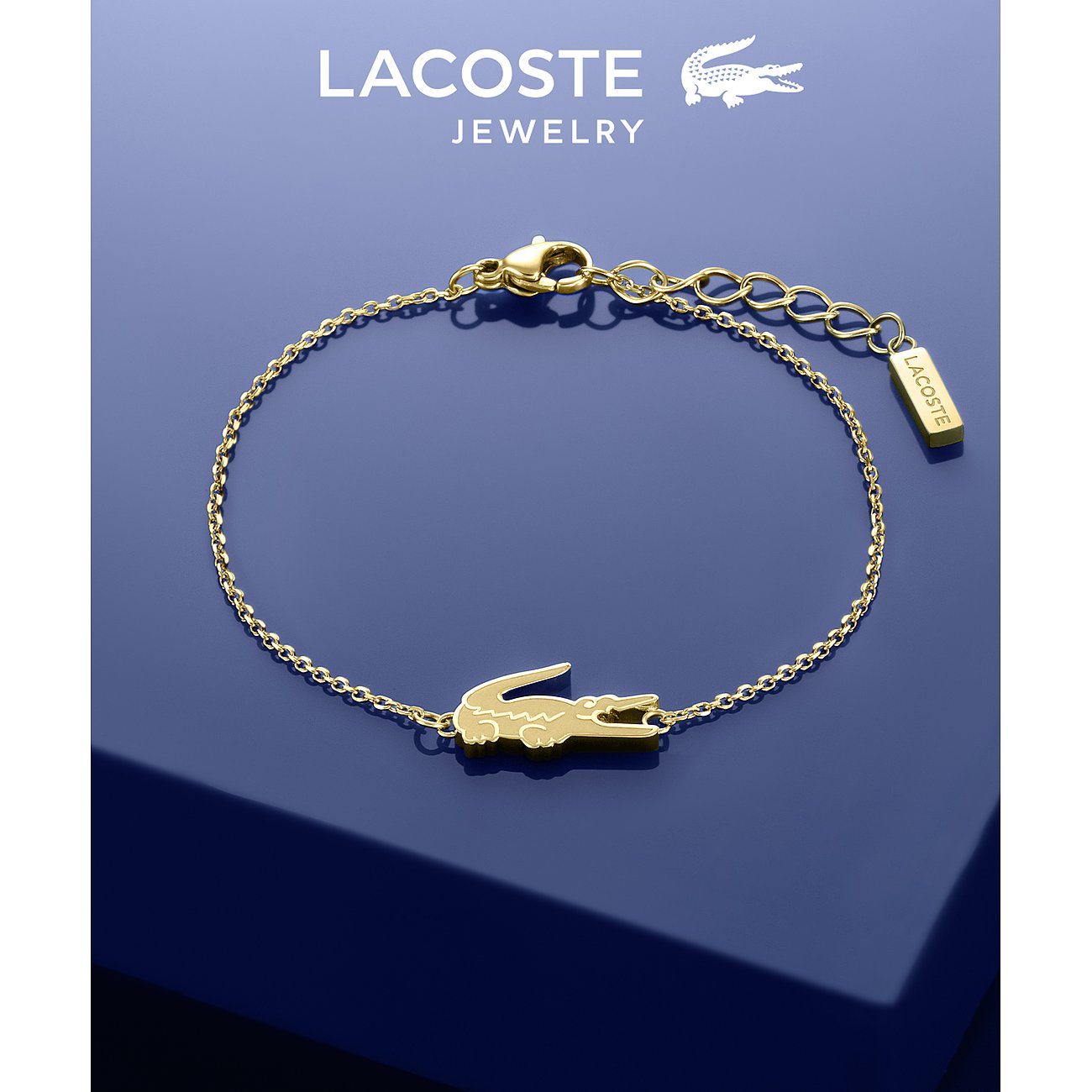 Lacoste armband van goudkleurig roestvrij staal met krokodilsymbool op marineblauwe achtergrond.