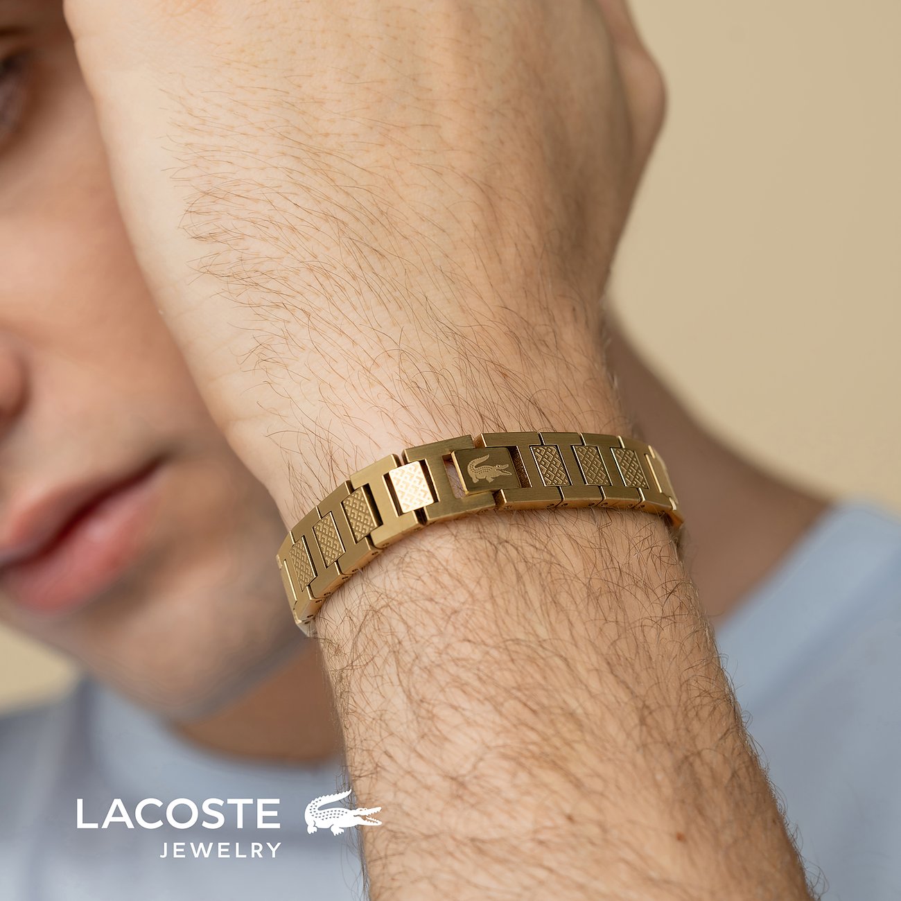 Een gouden Lacoste armband van roestvrij staal. Zichtbaar om de pols van een man op de voorgrond.
