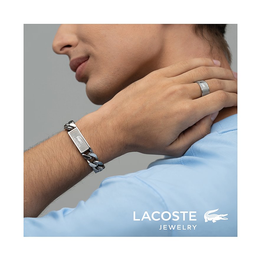 Close-up van man met Lacoste roestvrijstalen armband (brede ketting, gegraveerd logo), bijpassende ring en blauw poloshirt.