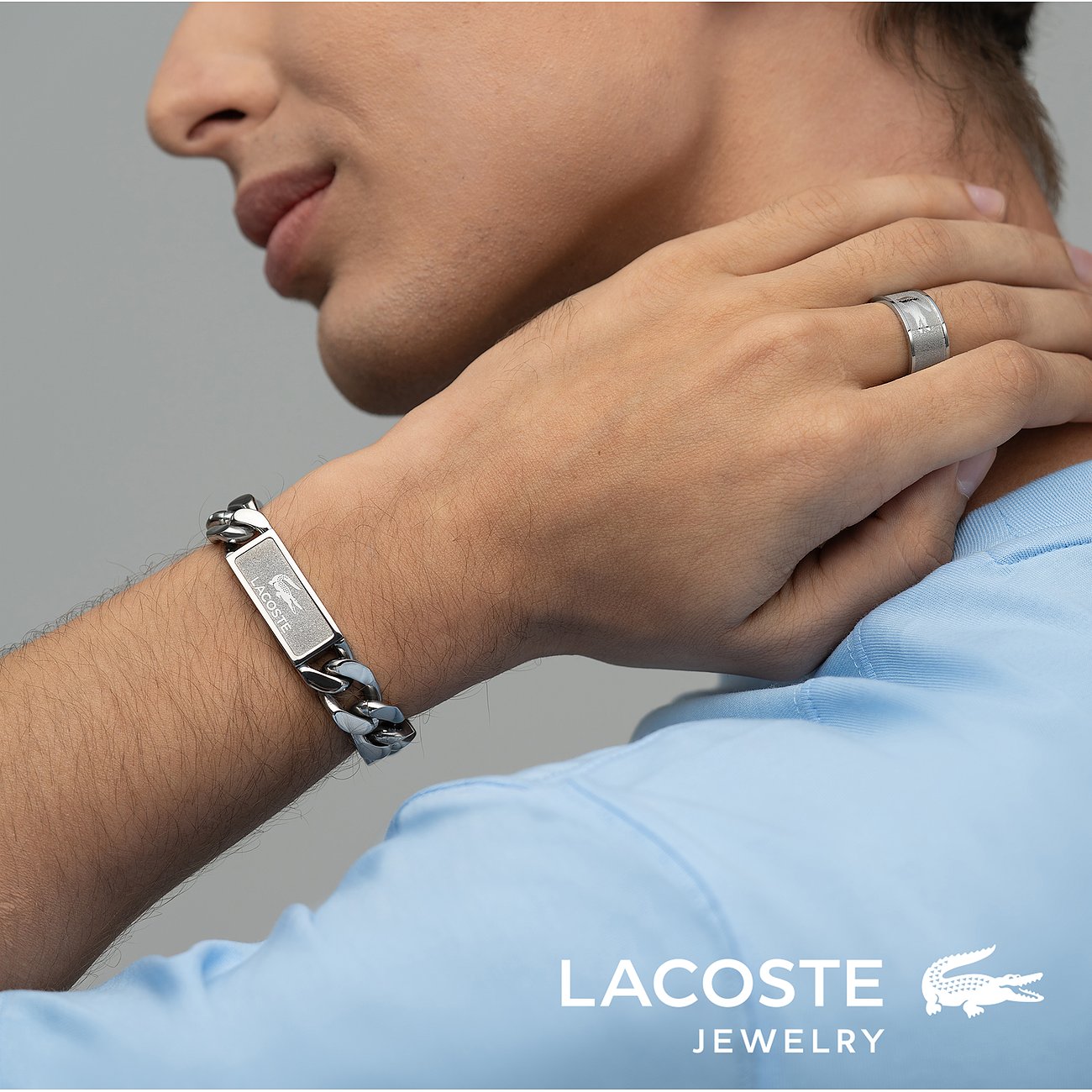 Close-up van man met Lacoste roestvrijstalen armband (brede ketting, gegraveerd logo), bijpassende ring en blauw poloshirt.