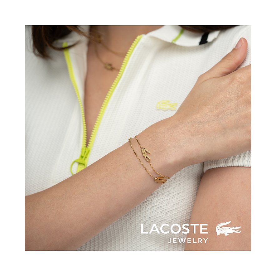 Een Lacoste armband van goudkleurig roestvrij staal met een krokodilbedel, gedragen om de pols van een vrouw.