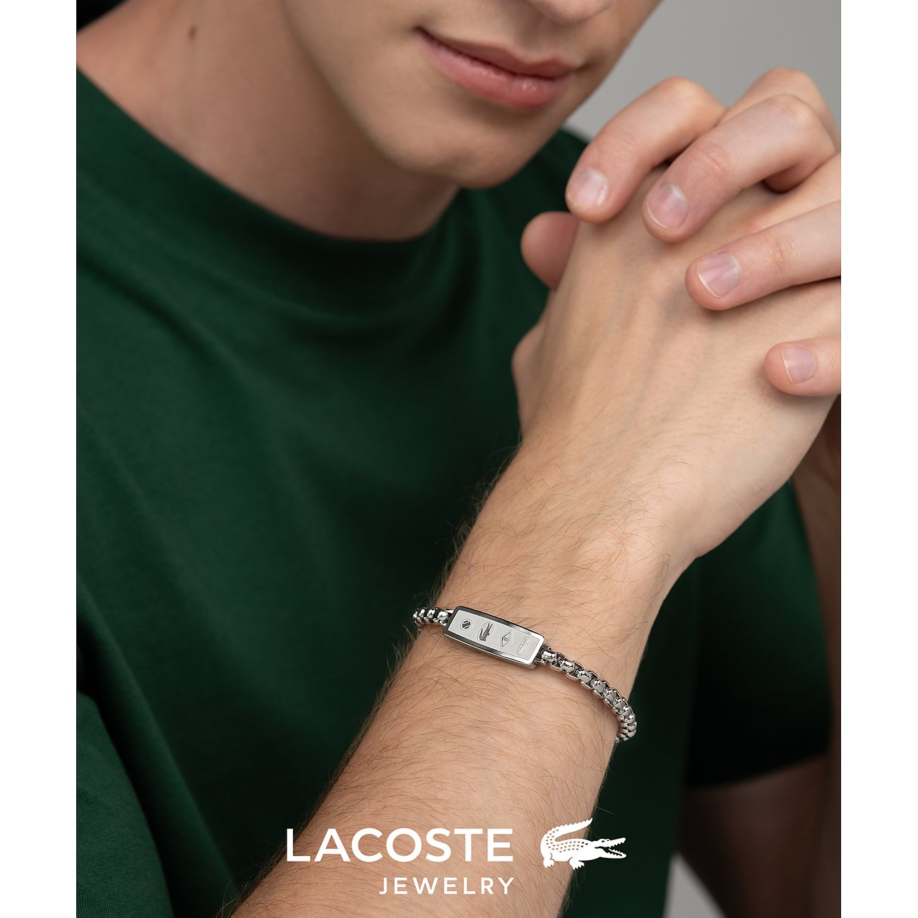 Close-up van man met zilverkleurige Lacoste roestvrijstalen schakelarmband met rechthoekig plaatje.