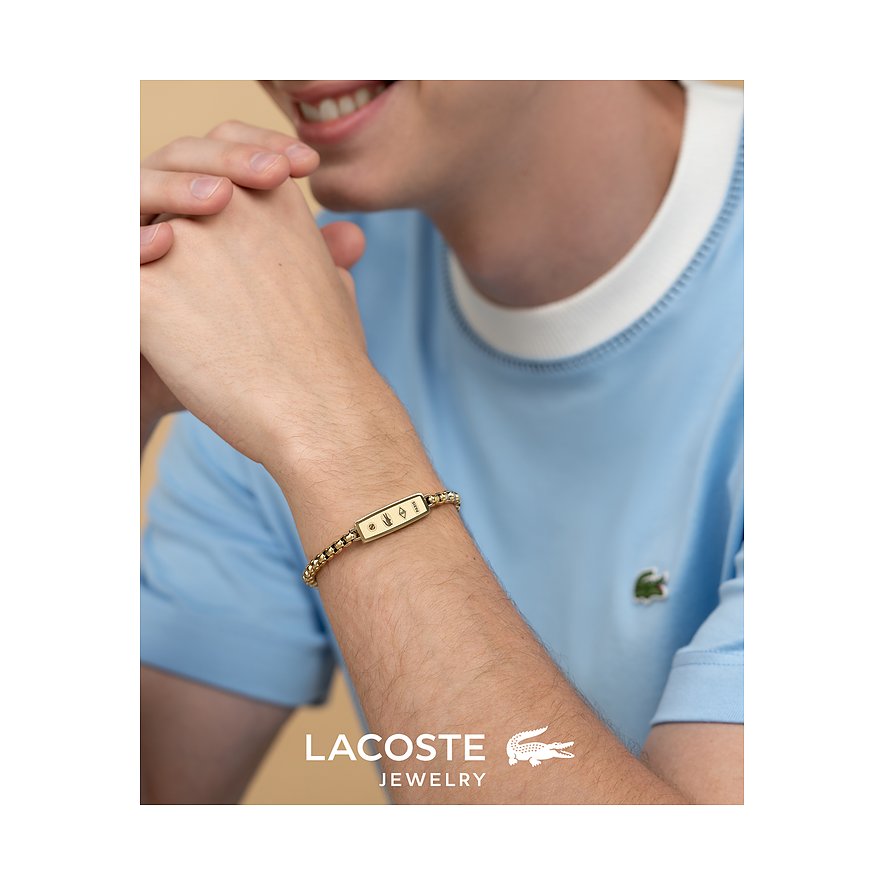 Nahaufnahme eines Lacoste Armschmucks aus goldfarbenem Edelstahl am Handgelenk eines Mannes mit hellblauem T-Shirt.
