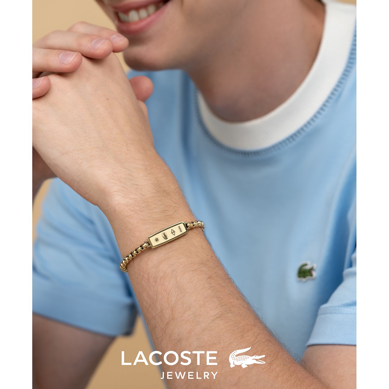 Nahaufnahme eines Lacoste Armschmucks aus goldfarbenem Edelstahl am Handgelenk eines Mannes mit hellblauem T-Shirt.