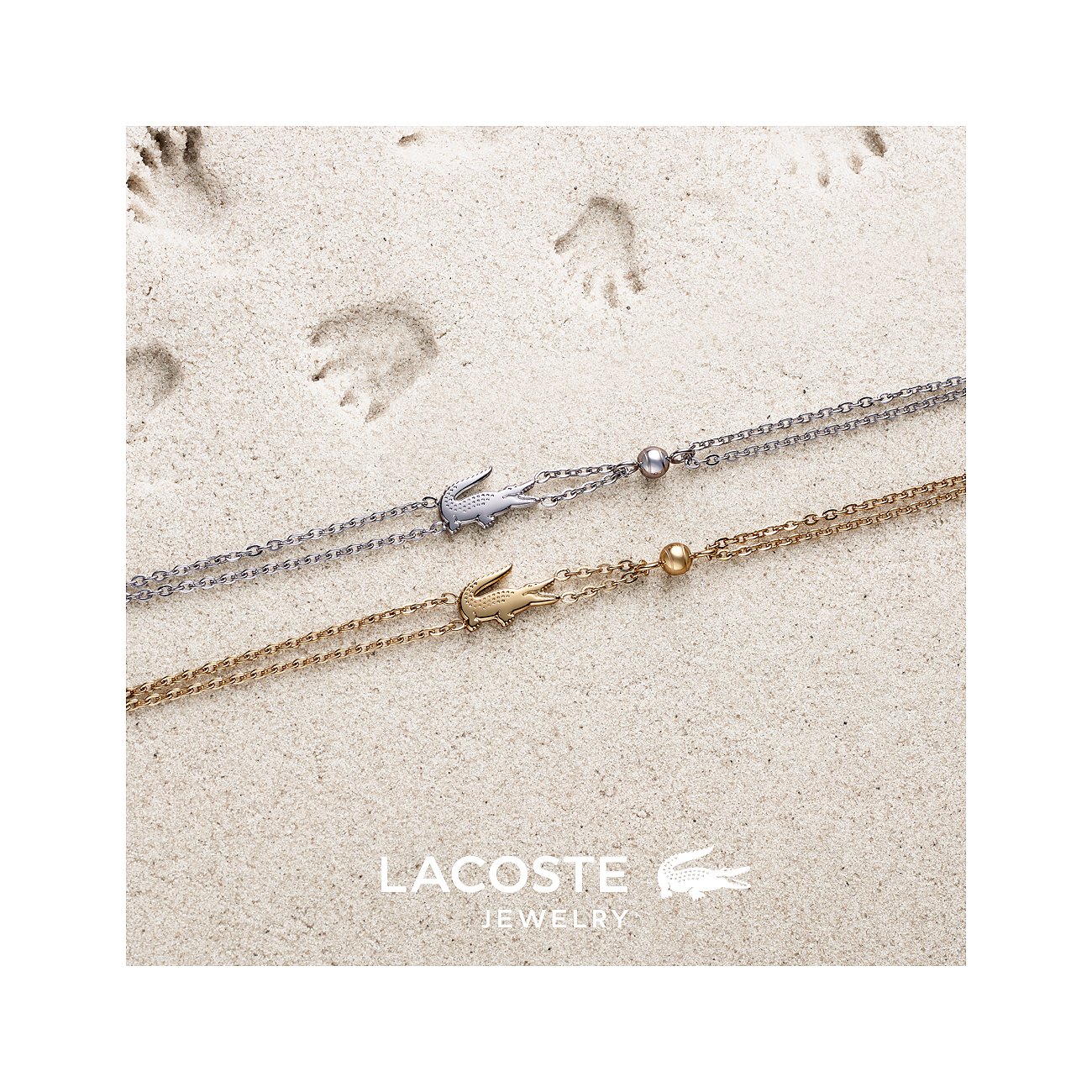 Twee Lacoste roestvrijstalen armbanden op zand. Eén is zilverkleurig, de andere goudkleurig.