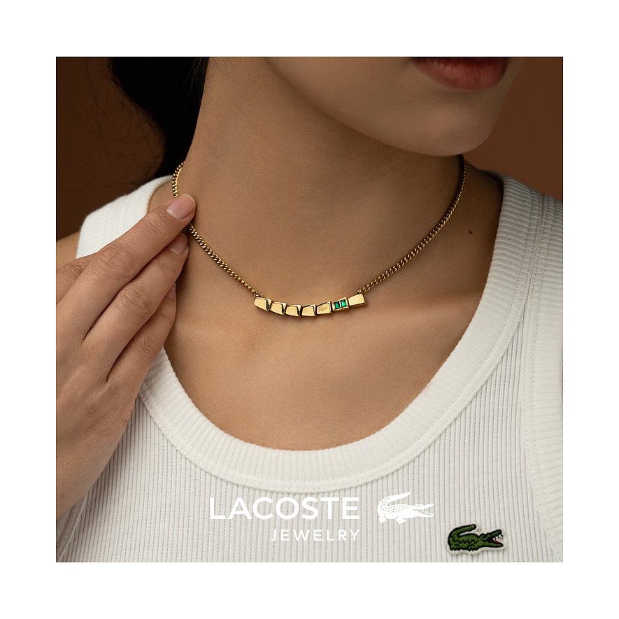 Een vrouw draagt een goudkleurige Lacoste roestvrijstalen ketting met rechthoekige hangers en een groene kristal.