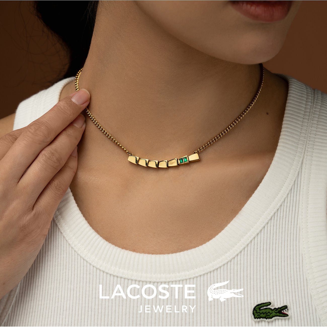 Een vrouw draagt een goudkleurige Lacoste roestvrijstalen ketting met rechthoekige hangers en een groene kristal.