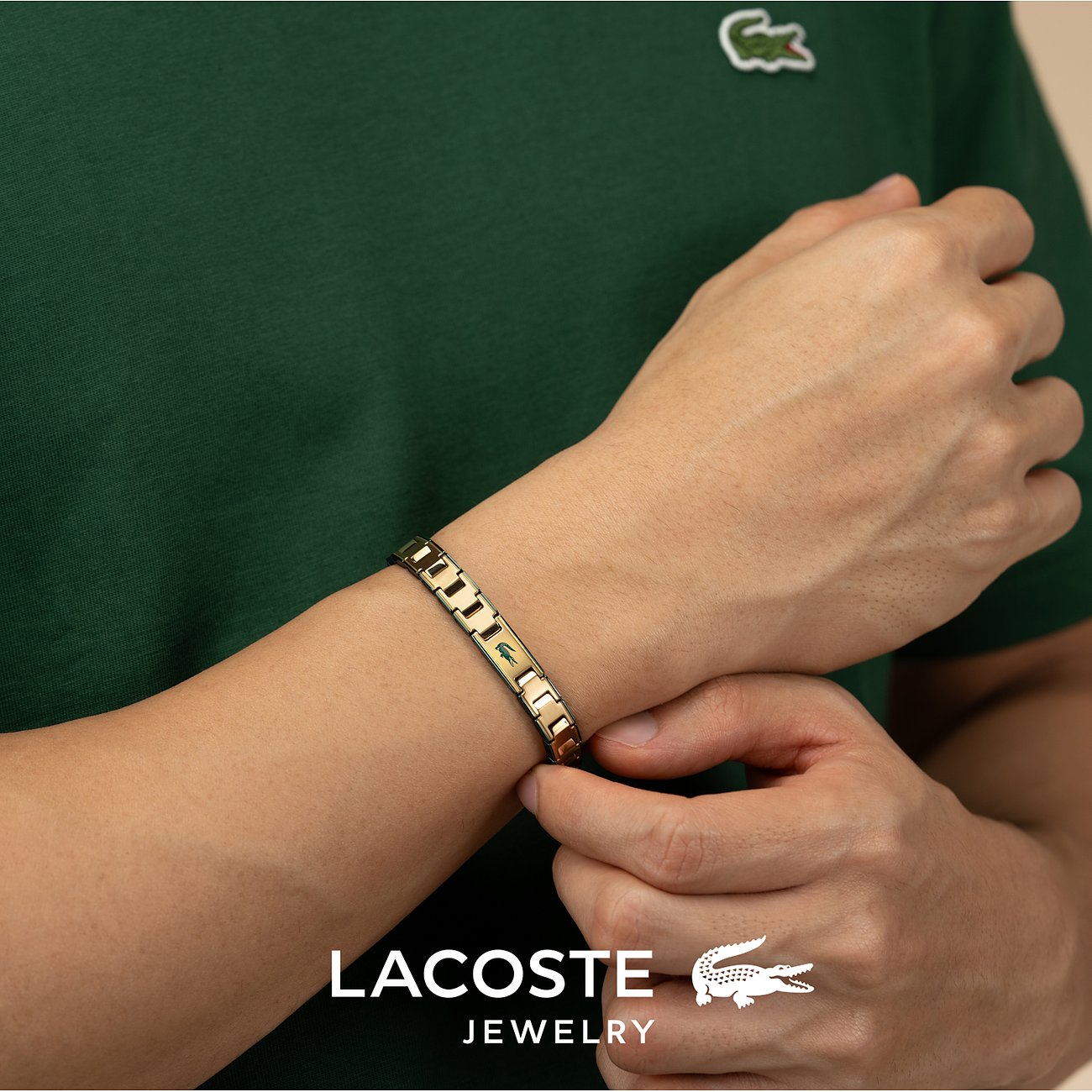 Close-up van een gouden Lacoste roestvrijstalen armband om de pols van een man met een groen shirt.