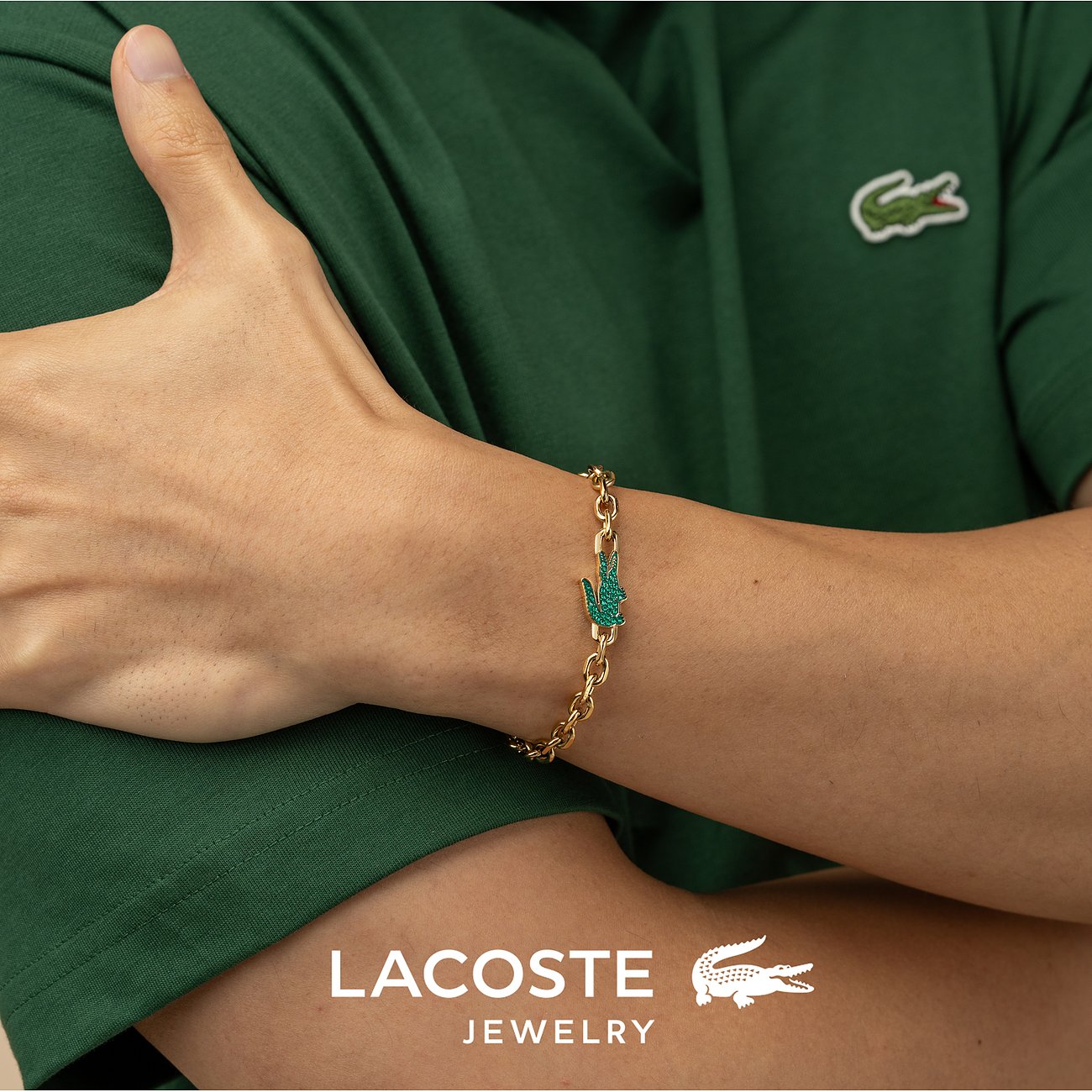 Am Handgelenk eines Models getragenes goldenes Lacoste Armband aus Edelstahl mit Krokodillogo aus grünem Kristall.